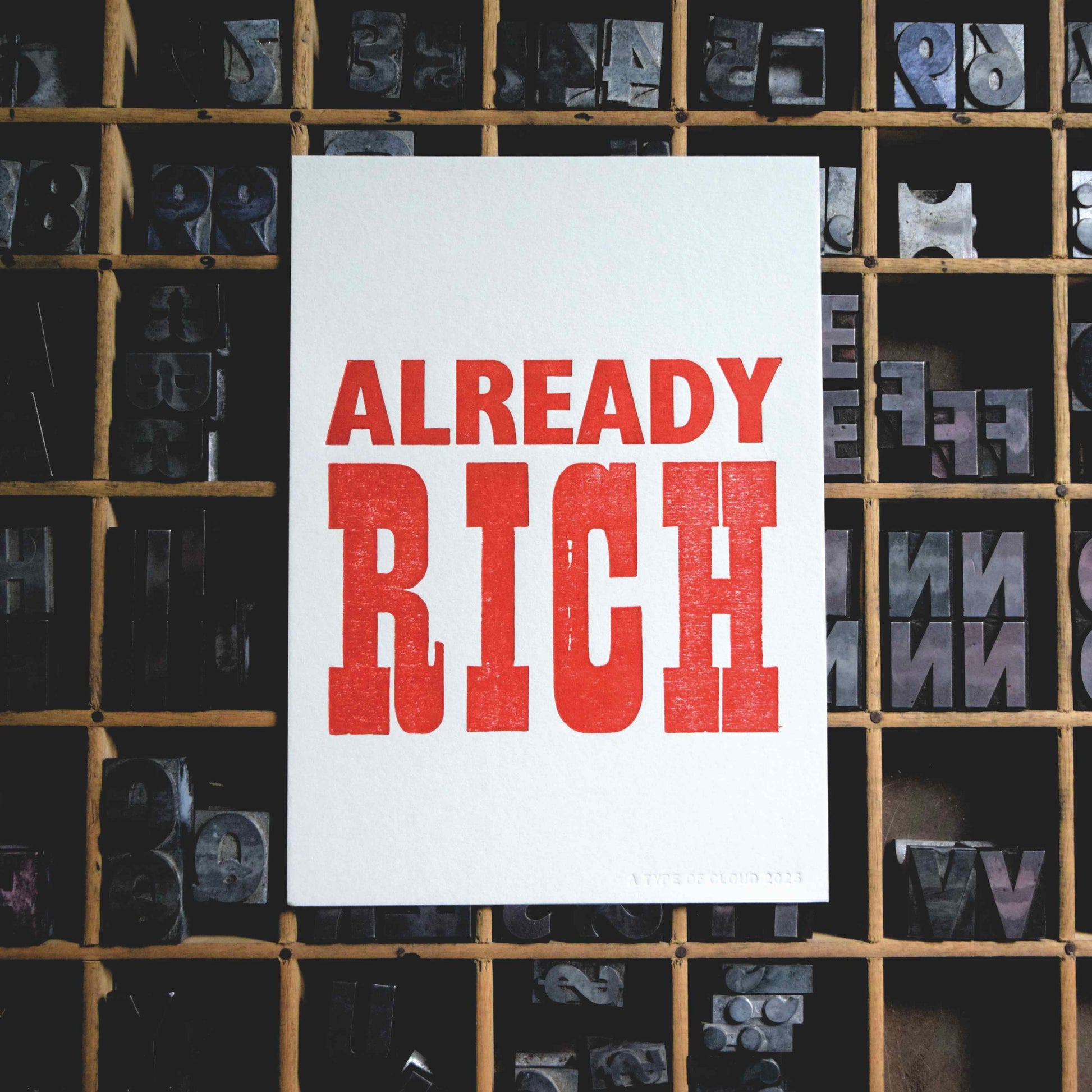 White card with 'ALREADY RICH' in red text against a background of letterpress printing blocks.