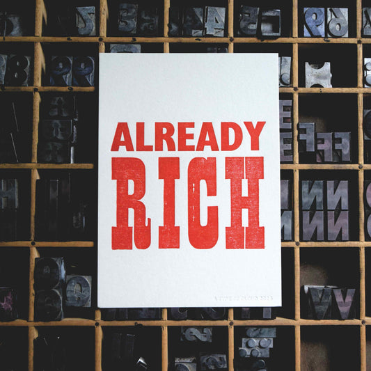 White card with 'ALREADY RICH' in red text against a background of letterpress printing blocks.